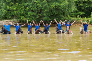 Image of Kelsey and Michael Gascon posing with the Horse Help Train-Off contestants in a body of water, accompanied by their horses. The scene captures a moment of unity, camaraderie, and equestrian adventure during the competition.