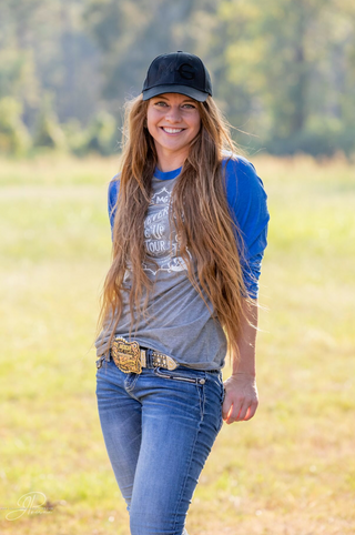 Kelsey Gascon elegantly models the MG cap paired with a stylish grey raglan adorned with blue sleeves featuring the Never Give Up tour logo, showcasing fashionable merchandise options.