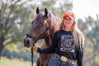 A Horse Help staff member confidently models the MG cap and a Horse Help long sleeve shirt featuring the Never Give Up tour logo, adding a touch of style and support to their ensemble.